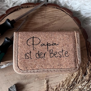 Könnte beinhalten: Ein Kartenhalter aus Kork mit dem Text "Papa ist der Beste" in schwarzer Schrift. Der Kartenhalter liegt auf einer Holzoberfläche mit zwei Schraubenziehern und dekorativen getrockneten Pflanzen.