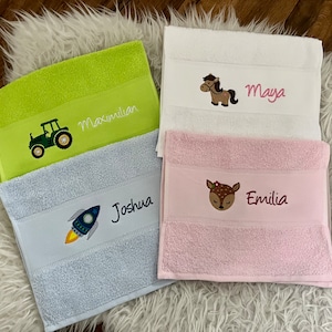 Könnte beinhalten: Vier bestickte Handtücher mit verschiedenen Designs und Namen. Ein grünes Handtuch mit einem Traktor und dem Namen Maximilian, ein blaues Handtuch mit einer Rakete und dem Namen Joshua, ein weißes Handtuch mit einem Pferd und dem Namen Maya und ein rosa Handtuch mit einem Reh und dem Namen Emilia.