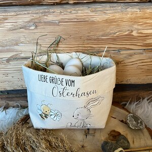 K&ouml;nnte beinhalten: Ein wei&szlig;er Canvas-Osterkorb mit einem Hasen- und Bienen-Design. Der Korb ist mit Heu und wei&szlig;en Eiern gef&uuml;llt. Der Text auf dem Korb lautet "Liebe Gr&uuml;&szlig;e vom Osterhasen".