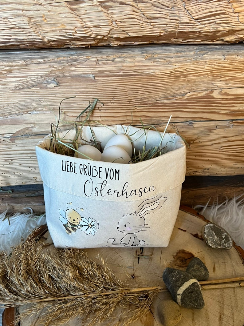 K&ouml;nnte beinhalten: Ein wei&szlig;er Canvas-Osterkorb mit einem Hasen- und Bienen-Design. Der Korb ist mit Heu und wei&szlig;en Eiern gef&uuml;llt. Der Text auf dem Korb lautet "Liebe Gr&uuml;&szlig;e vom Osterhasen".
