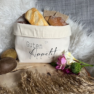 Op de afbeelding: Een witte canvas broodmand met de tekst "Guten Appetit" en een hartje. De mand is gevuld met broodjes en broden. De mand staat op een houten oppervlak met een pluche wit oppervlak op de achtergrond.