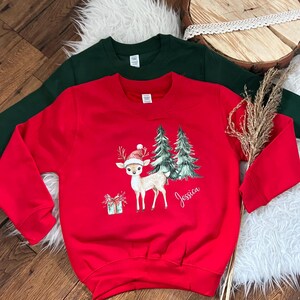 Könnte beinhalten: Rotes Weihnachts-Sweatshirt mit einem Hirsch mit Weihnachtsmütze, einem Geschenk und zwei Weihnachtsbäumen. Der Name "Jessica" ist in Schreibschrift geschrieben. Ein dunkelgrünes Sweatshirt befindet sich hinter dem roten.