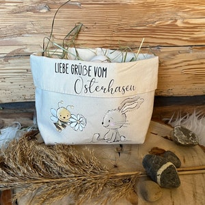 K&ouml;nnte beinhalten: Ein wei&szlig;er Canvas-Osterkorb mit einem Hasen- und Bienen-Design. Der Korb ist mit Stroh gef&uuml;llt und hat den Text "Liebe Gr&uuml;&szlig;e vom Osterhasen" auf der Vorderseite.