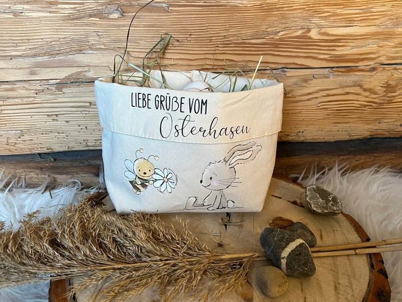 K&ouml;nnte beinhalten: Ein wei&szlig;er Canvas-Osterkorb mit einem Hasen- und Bienen-Design. Der Korb ist mit Stroh gef&uuml;llt und hat den Text "Liebe Gr&uuml;&szlig;e vom Osterhasen" auf der Vorderseite.