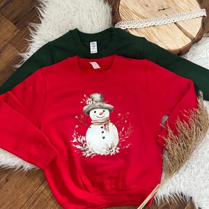 Könnte beinhalten: Ein rotes Sweatshirt mit einem Schneemann-Design, das einen grauen Hut, Schal und Blumendetails aufweist. Ein dunkelgrünes Sweatshirt ist dahinter zu sehen. Die Sweatshirts liegen auf einer weißen, flauschigen Oberfläche.