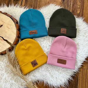 Könnte beinhalten: Vier verschiedene farbige Strickmützen mit Lederpatches. Die Mützen sind türkis, olivgrün, senfgelb und rosa. Die Patches haben folgenden Text: "Be happy", "Be wild & wonderful", "Wild one" und "Lieblings Mensch".