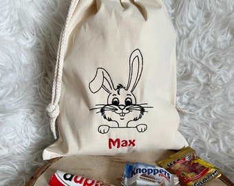 May include: A white canvas drawstring bag with an embroidered bunny rabbit and the name "Max". The bag is filled with various candy bars, including a chocolate bar with the name "aupi" printed on it.