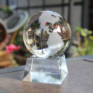 May include: A clear glass globe with a frosted etched world map sits on a crystal base. The base is engraved with the text "WYNDHAM Desert Blue", "Zina Kadmiri", "Dec 2020", and "Rep".