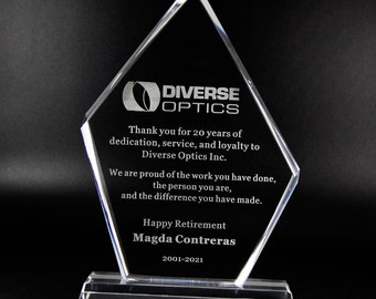 Op de afbeelding: Een heldere, ruitvormige glazen award met het Diverse Optics logo en gegraveerde tekst die Magda Contreras bedankt voor 20 jaar dienst, inclusief de data 2001-2021, gemonteerd op een rechthoekige, heldere glazen voet.