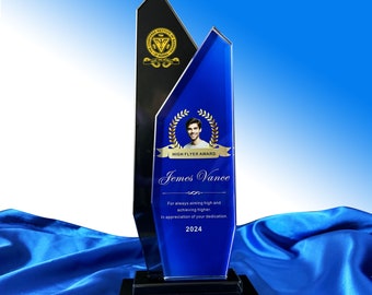 Op de afbeelding: Een High Flyer Award trofee met een zwart en blauw glazen ontwerp. Het blauwe gedeelte bevat een foto, een lauwerkrans en de naam "James Vance". De tekst luidt: "Voor altijd hoog mikken en hoger presteren. Ter waardering van uw toewijding. 2024."