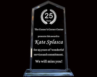 Op de afbeelding: Een heldere glazen award-plaquette met een puntige bovenkant en een rechthoekige voet. Het oppervlak bevat een witte lauwerkransafbeelding met de tekst 25 Years, The Cosner's Corner Center, Kate Splasca en de boodschap for 25 years of wonderful service and commitment. We will miss you!