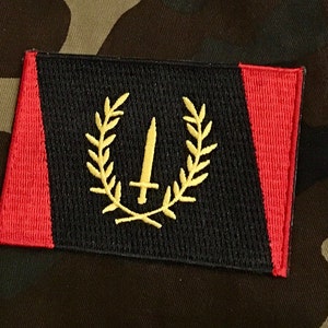 Può includere: Toppa ricamata con centro nero, pannelli laterali rossi e un emblema dorato. L'emblema presenta una spada all'interno di una corona d'alloro. La toppa è rettangolare e ha uno sfondo mimetico.