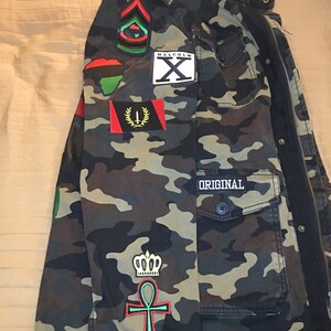 Peut inclure: Veste de camouflage avec divers patchs, dont un patch de l'Afrique continentale rouge, noir et vert, un patch "Malcolm X" noir et blanc, un patch vert, noir et rouge avec un symbole, un patch de drapeau rouge, noir et or, un patch vert et rouge avec une couronne et un symbole ankh, et un patch noir avec le mot "ORIGINAL".