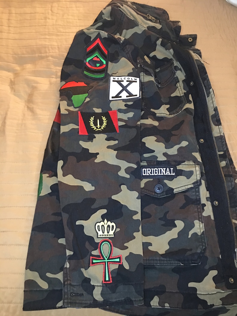 Peut inclure: Veste de camouflage avec divers patchs, dont un patch de l'Afrique continentale rouge, noir et vert, un patch "Malcolm X" noir et blanc, un patch vert, noir et rouge avec un symbole, un patch de drapeau rouge, noir et or, un patch vert et rouge avec une couronne et un symbole ankh, et un patch noir avec le mot "ORIGINAL".