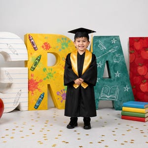 May include: A young child in a graduation gown and cap stands in front of large, colorful letters spelling "GRAD." The letters are decorated with school-themed elements like crayons, apples, and a chalkboard.