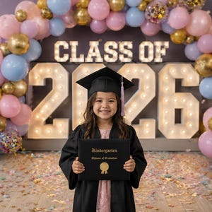 May include: A child in a graduation gown and cap smiles, holding a diploma in front of a backdrop with "CLASS OF 2026". The background has a balloon arch in gold, pink, and blue. Confetti is scattered on the floor.