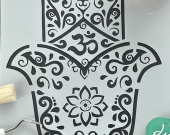 Può includere: Uno stencil in bianco e nero di una mano di hamsa con dettagli intricati e un simbolo Om al centro. Lo stencil è su uno sfondo grigio e ci sono pennelli e una piccola pietra dipinta nelle vicinanze.