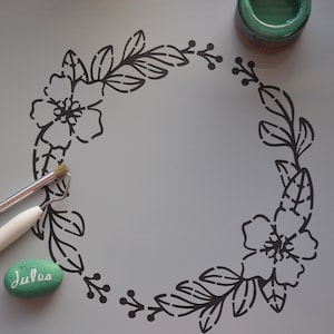May include: A black floral wreath stencil with a stone painted with the word "Jules".