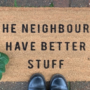 May include: A brown coir doormat with the text "The neighbours have better stuff" printed in black. A pair of black shoes are standing on the mat.