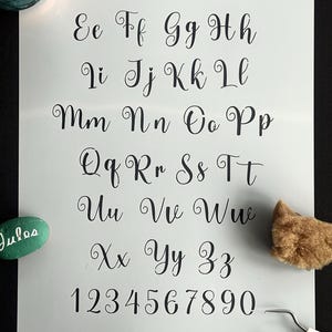 Pode incluir: Uma folha branca exibe um alfabeto completo e números em uma fonte cursiva preta elegante. A imagem inclui uma pedra verde com o nome "Jules" escrito, juntamente com materiais de arte, como um pincel e uma esponja.