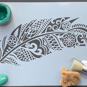 Può includere: Uno stencil con un designo di piuma dettagliato con motivi e volute intricati. Lo stencil è su uno sfondo bianco ed è circondato da materiali per l'artigianato, tra cui un piccolo barattolo di vernice verde, una spugna e un pennello.