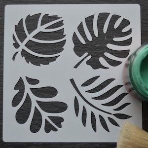 May include: A white plastic stencil featuring four different leaf designs. The stencil is on a black surface with a green paint jar and a paint brush.