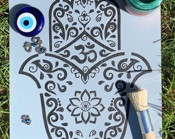 Può includere: Uno stencil di una mano di hamsa con dettagli intricati, tra cui un simbolo Om e un motivo floreale. Lo stencil è su uno sfondo blu chiaro ed è circondato da ciondoli blu di occhio turco e un pennello.