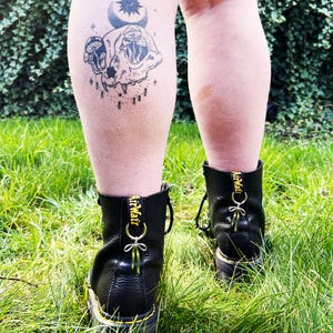 May include: A person wearing black leather boots with yellow stitching and the text "AirWair" on the back. The person has a black and white tattoo of a skull with mushrooms on their lower leg.