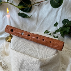 May include: A brown wooden flute with a textured, bark-like finish. The flute features several finger holes and a mouthpiece. It is placed on a beige fabric surface, with a lit candle and foliage in the background.
