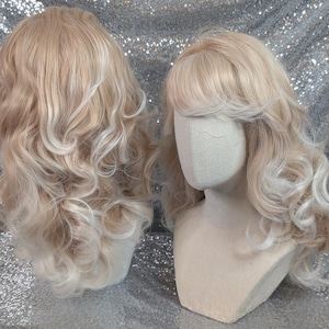 May include: Two blonde wigs displayed on mannequin heads. The wigs feature long, wavy hair with a mix of light blonde and platinum white highlights. One wig is shown from the back, while the other is shown from the front with bangs.