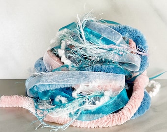 May include: A bundle of assorted decorative ribbons and yarn in shades of blue, pink, and white. The collection includes textured chenille yarn, sheer organza ribbons, and frayed metallic fibers, all intertwined to create a multi-textured cluster.