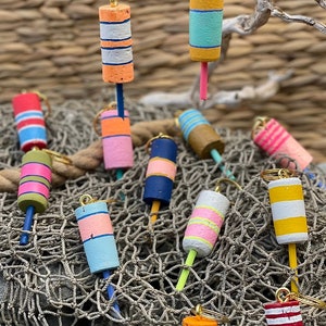May include: A collection of colorful, hand-painted cork buoys with gold key rings. The buoys are painted in various patterns with stripes and colors like red, white, blue, yellow, pink, and green. The buoys are displayed on a brown, woven net.