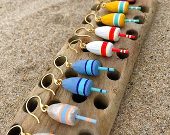 May include: A collection of colorful buoy-shaped earrings with gold-colored hooks. The earrings are arranged on a piece of driftwood, with each buoy painted in a different color scheme, including pink, yellow, white, and blue, with orange and blue stripes.