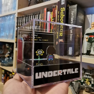May include: A clear acrylic cube display case with a black base featuring the text "UNDERTALE". Inside the case is a pixelated image of a yellow flower with a smiling face and a character from the video game "Undertale". The text "Howdy! I am FLOWEY.  FLOWEY the FLOWER!" is displayed in a black speech bubble.