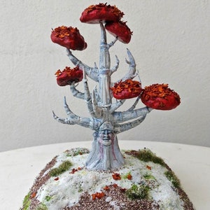 May include: A white, skeletal tree with a face carved into the trunk. The tree has red, leaf-covered branches and is set on a snowy base with green moss.