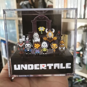 Könnte beinhalten: Ein durchsichtiger Würfel-Präsentationskasten aus Kunststoff mit schwarzem Sockel, auf dem der Text "UNDERTALE" in weißen Pixelbuchstaben mit einem roten Herzen steht. Im Inneren des Kastens befinden sich 10 kleine Pixelfiguren aus dem Videospiel "Undertale".