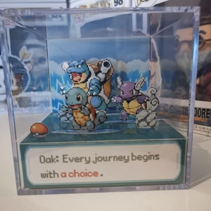 Puede incluir: Una caja de presentación de cubo de plástico transparente con una escena emergente en 3D de personajes de Pokémon. La escena presenta a Squirtle, Wartortle y Blastoise. La base de la caja de presentación tiene un fondo verde con el texto "Oak: Every journey begins with a choice."