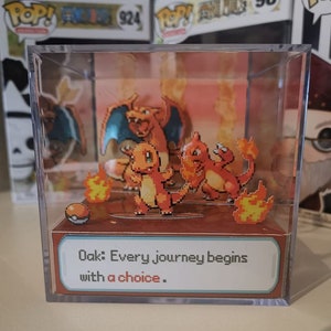 May include: A clear plastic cube display case with a 3D pixel art scene of three Pokemon characters: Charmander, Charmeleon, and a Charizard. The scene is set against a background of orange flames. The quote "Oak: Every journey begins with a choice." is printed on a white card at the bottom of the display case.