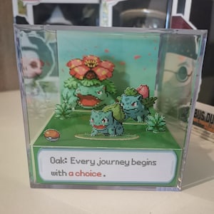 Könnte beinhalten: Ein durchsichtiger Würfel-Präsentationskoffer aus Kunststoff mit einer 3D-Pop-up-Szene des Pokémon Bisasam. Die Szene zeigt drei Bisasam-Figuren, einen Pokéball und einen grünen Hintergrund mit rosa Blüten. Der Text "Oak: Every journey begins with a choice." ist auf einer weißen Karte am Boden des Präsentationskoffers gedruckt.