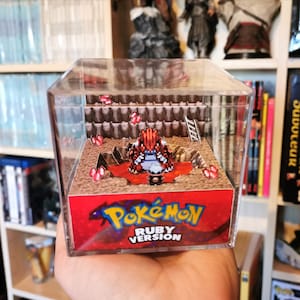 Può includere: Una teca di esposizione cubica in plastica trasparente con un diorama di carta 3D al suo interno. Il diorama rappresenta un personaggio Pokémon rosso e blu, probabilmente un Pokémon iniziale, in piedi in una fossa di lava rossa. Il diorama è etichettato "Pokémon Versione Rubino".