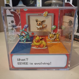 Puede incluir: Una caja de presentación de cubo de plástico transparente con una escena de Pokémon colorida en su interior. La escena presenta un Eevee pixelado que evoluciona a sus diferentes formas. El texto "What? EEVEE is evolving!" está impreso en la parte inferior de la caja de presentación.