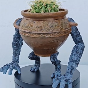 Può includere: Un vaso in terracotta a forma di creatura che cammina con un cactus che cresce al suo interno. Il vaso ha una finitura marrone e nera e si trova su una base circolare nera.