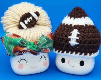 May include: Two white ceramic mugs with cute faces. One mug has a brown and white crocheted football hat. The other mug has a brown and white yarn bun with a brown and white football on top. Both mugs have a green and brown patterned ribbon around the top.