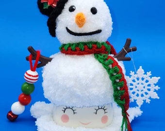 May include: A white snowman with a black hat, red scarf, and green and red scarf. The snowman has a red and white beaded garland attached to its arm. The snowman is sitting on a white mug with a smiling face. A white snowflake is in the background.