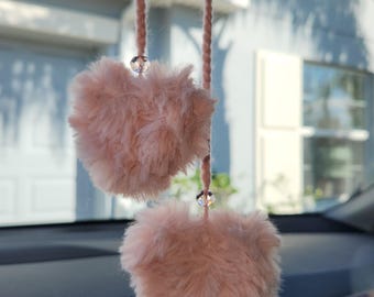May include: Two pink fuzzy heart-shaped car air fresheners hanging from the rearview mirror of a car. The fresheners are attached to strings with clear crystal beads.