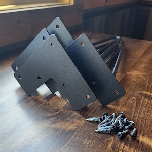 May include: Three black metal table leg brackets with four screw holes each, and a pile of silver screws on a dark brown wooden surface.