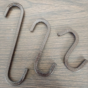 May include: Three metal S-shaped hooks in a dark brown finish. The hooks are different sizes and are arranged in a row on a wooden surface.