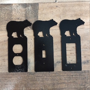 May include: Three black metal light switch and outlet covers with a bear silhouette design. The covers are for a single light switch, a double light switch, and a single outlet.