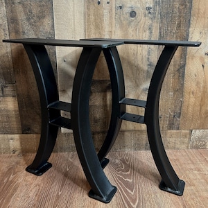 May include: Two black metal table legs with a curved design. The legs have a flat top and base, with a horizontal support bar. The legs are made of a sturdy metal and are designed to support a tabletop. The legs are shown against a wood background.