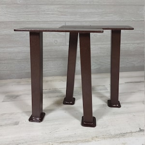 May include: Three metal table legs in a dark brown color. Two legs are in a triangular shape and one leg is a single post. The legs are made of metal and have a textured finish.